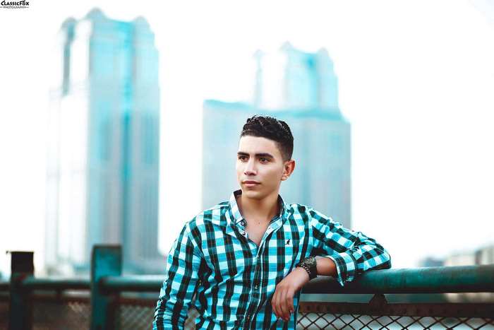 Young man in plaid shirt leaning on fence with buildings