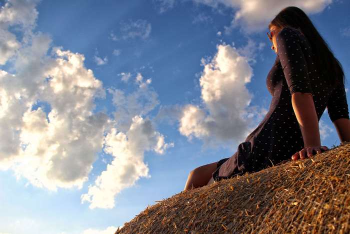 Woman sitting on hay bale under cloudy sky