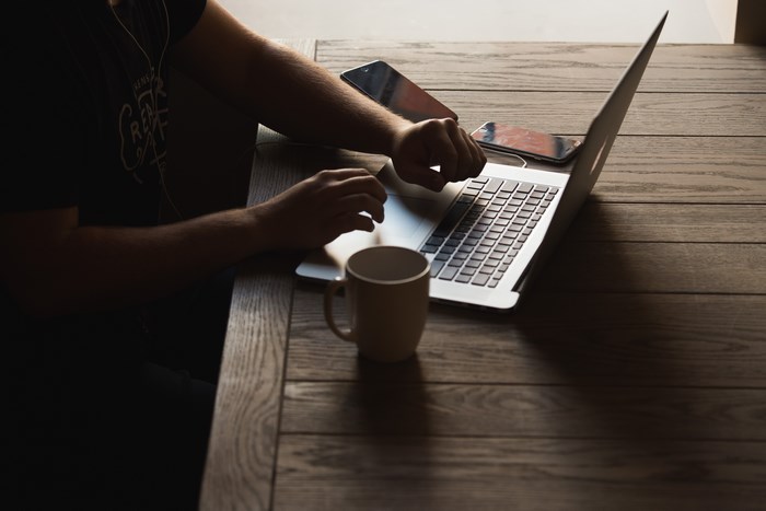 Person typing on a MacBook with a mug and phones