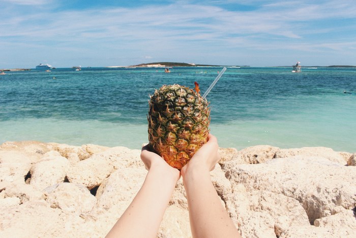 Hands holding a pineapple cocktail on a rocky beach with the ocean in the background