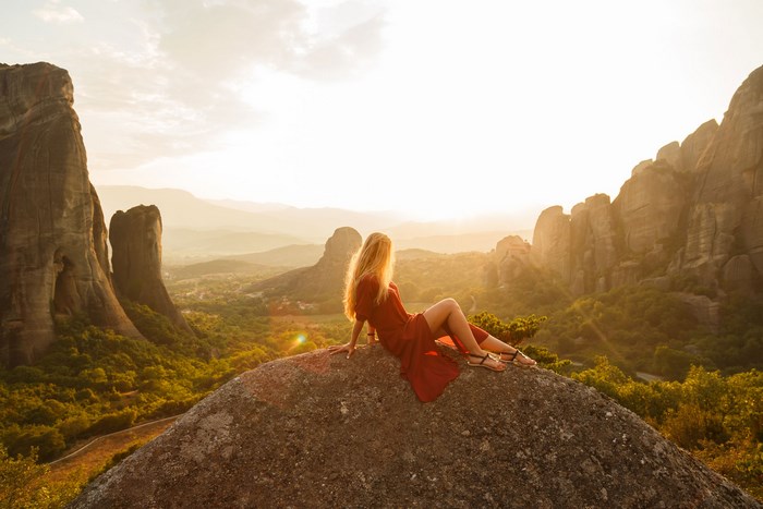 Woman in red dress sitting on boulder at sunset in Meteora