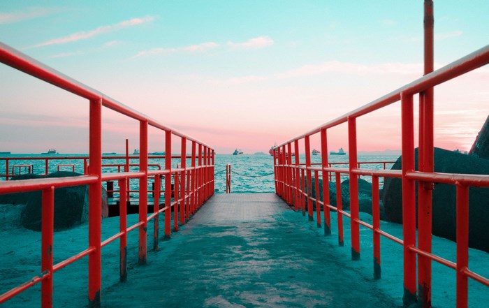 Pier with red railings at sunset with ships in the distance
