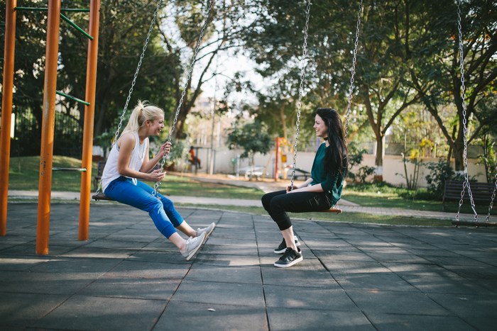 Two women talking and laughing on swings in a park