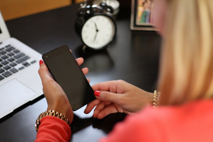 Woman holding smartphone at desk with laptop and clock
