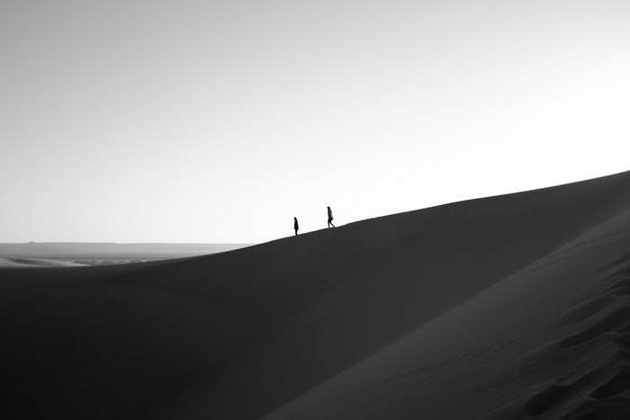 Two people walk across a vast sand dune