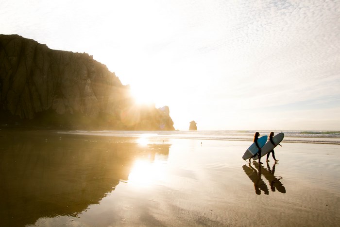 Two surfers walk along a beach at sunset with a cliff in the background.