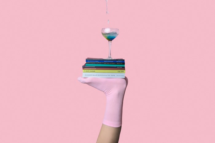A hand in a pink sock balances books and a cocktail glass