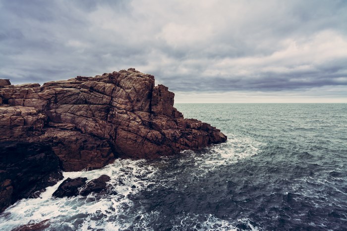Brown rocky cliff meets the ocean under a cloudy sky