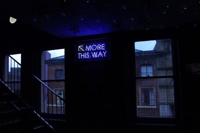Blue neon sign "MORE THIS WAY" in a dark room