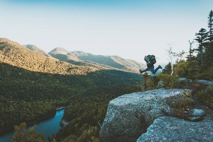 Hiker jumps off a rocky cliff overlooking a forest valley