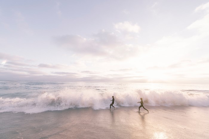 Two people in wetsuits running on a beach with waves