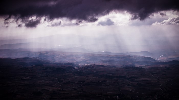 Sunbeams pierce through dark storm clouds over a mountainous landscape.