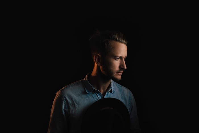 Man in denim shirt holding a hat in lowkey studio portrait