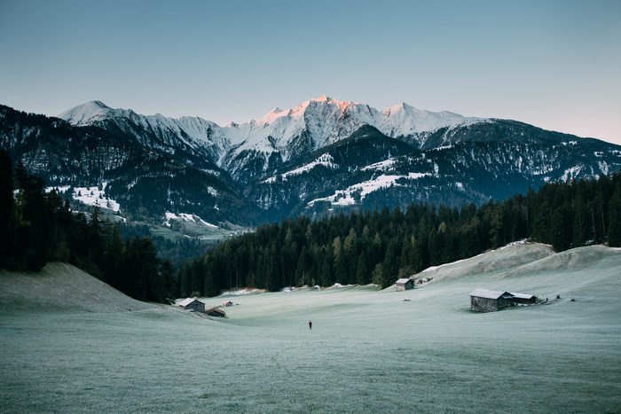 Lone person walks through a frosty field towards snow-capped mountains at sunrise