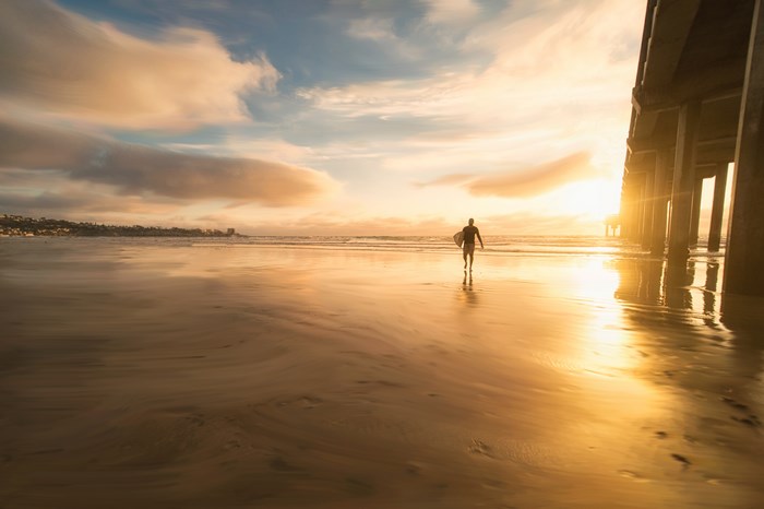 Surfer walking on beach at sunset under pier