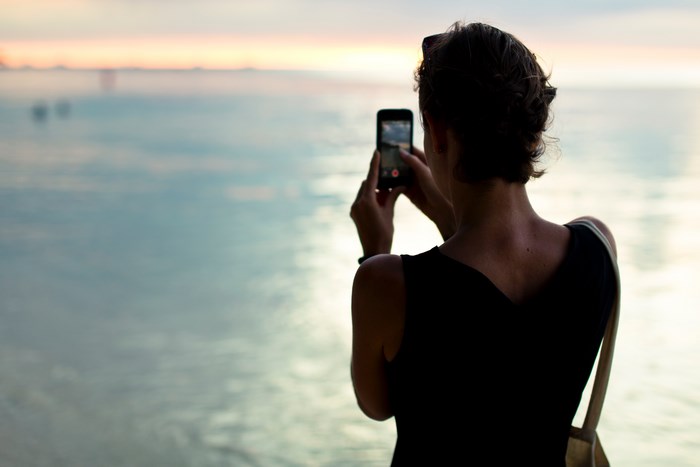 Woman taking a picture of the ocean at sunset with her phone