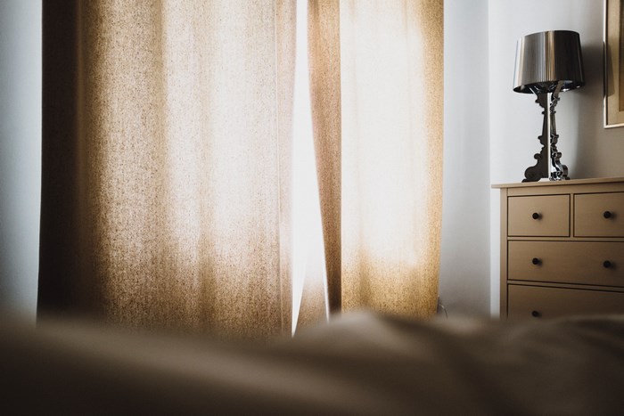 Sunlight through curtains in a bedroom with a dresser and lamp