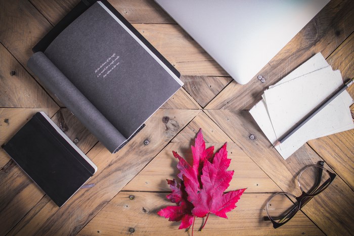 Flat lay of a laptop, books, glasses, and a red maple leaf on a wooden table