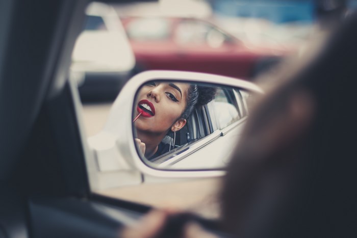 Woman applying red lipstick in car wing mirror