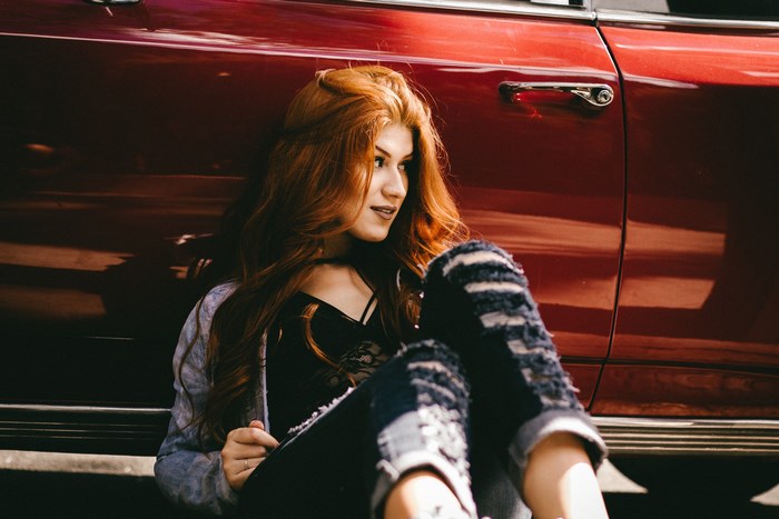 Redhead woman in ripped jeans sitting by a red car