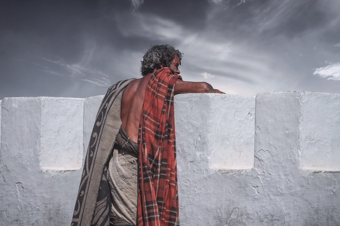 Man in blankets leans on a white wall under a cloudy sky