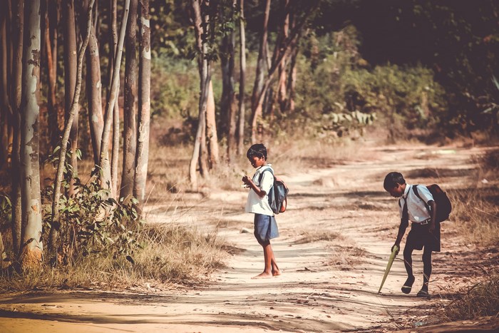 Two barefoot school children with backpacks on a dirt path