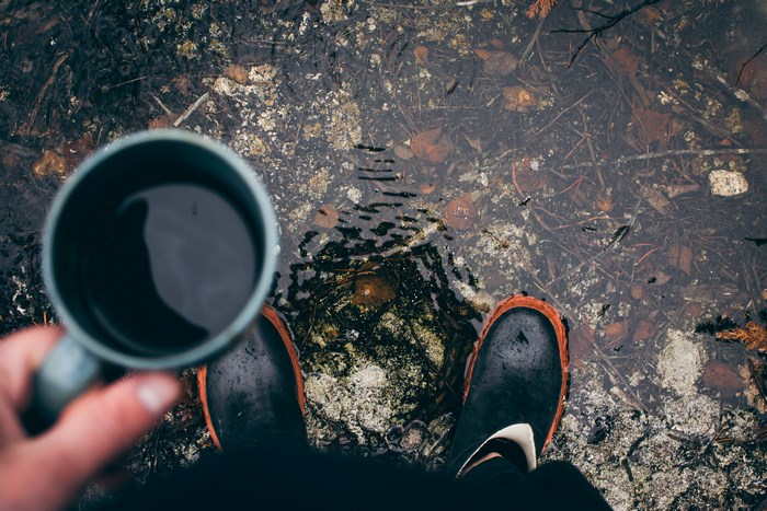 Person holding coffee cup standing in puddle with boots