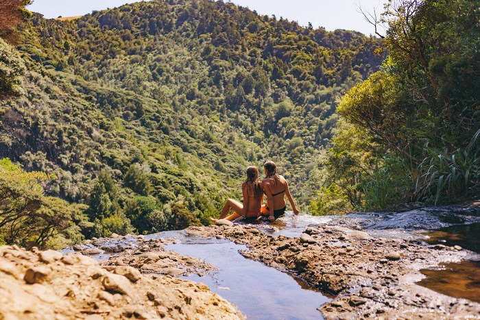 Two women sitting at the edge of a waterfall overlooking a valley