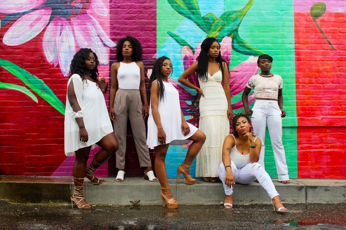 Six women in white outfits pose in front of a mural