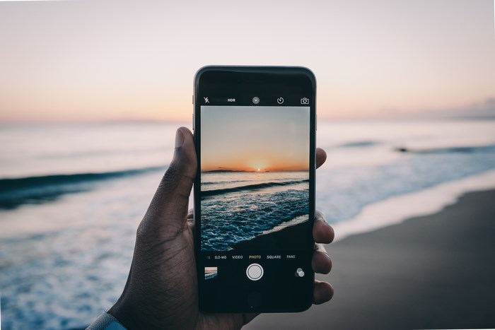 Hand holding iPhone taking a photo of a sunset over the sea