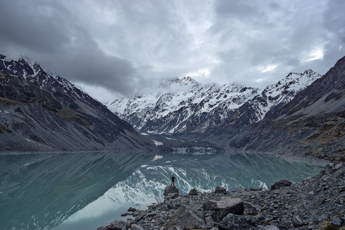 Person standing on rocks by a lake with snow-capped mountains