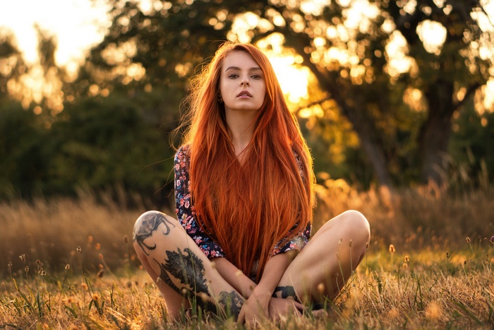 Woman with long red hair and tattoos sitting in a field