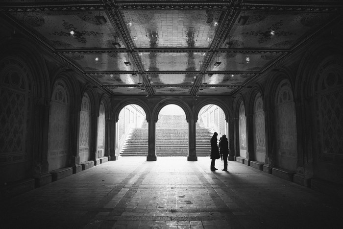 Two people stand in a grand arched hallway with stairs