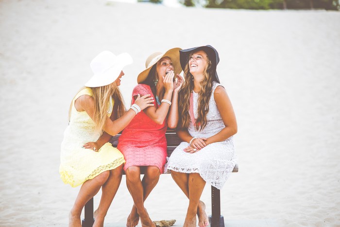 Three women in sunhats laughing on a bench at the beach