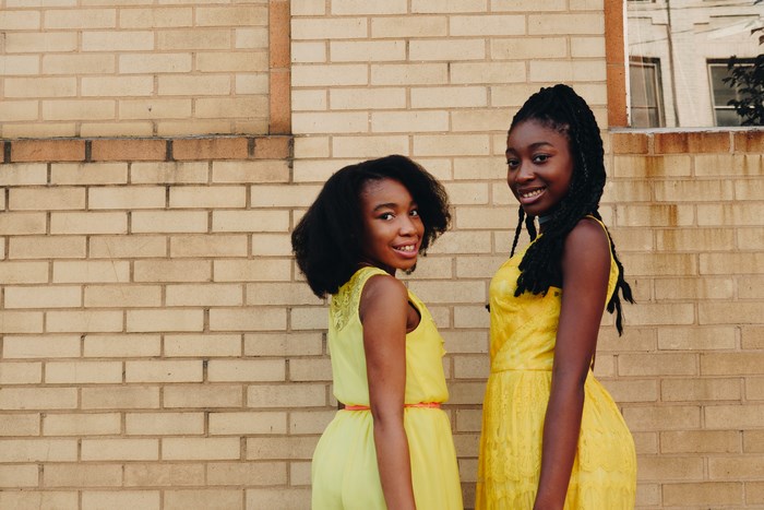 Two Smiling Black Girls in Yellow Dresses by Brick Wall