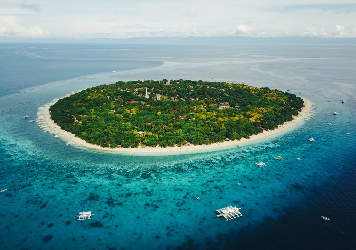 Aerial view of a circular island with lush green trees and white sand beaches surrounded by clear blue ocean water with boats.