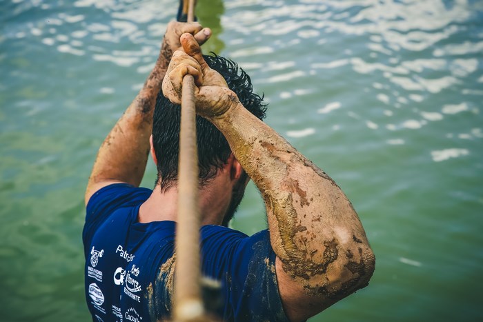 Man covered in mud climbing a rope over water