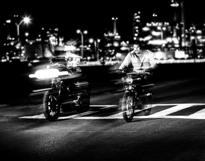 Two motorcyclists riding at night with city lights in background