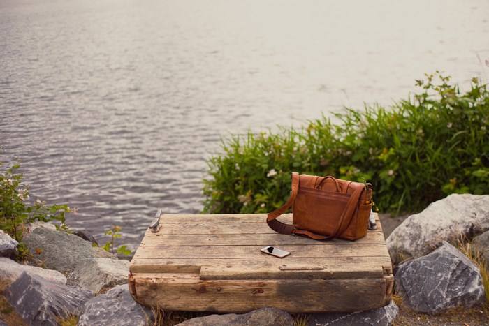 Brown leather satchel and phone on wooden surface by water