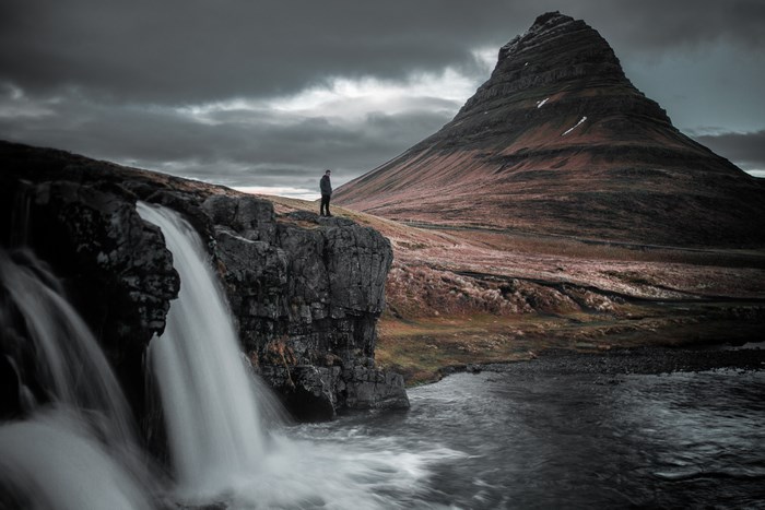 Man standing on cliff overlooking Kirkjufell waterfall and mountain in Iceland