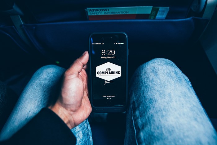 Man holding iPhone with "Stop Complaining" wallpaper on airplane