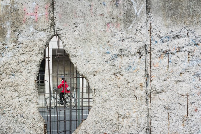 Woman in red coat cycling through a hole in the Berlin Wall