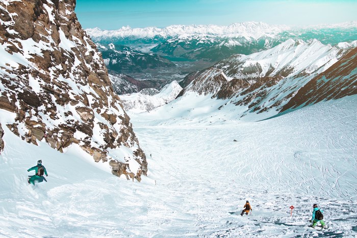 Skiers descend a snowy mountain slope with distant peaks