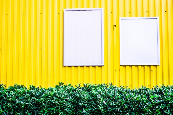 Yellow corrugated wall with two blank white frames and green hedge