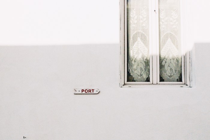 White wall with a sign pointing to the port and a window with lace curtains