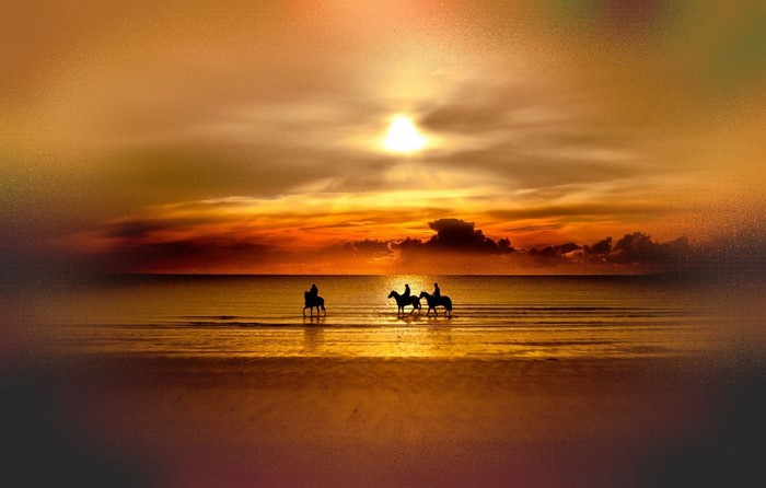 Three people on horseback at sunset over the water