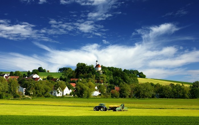 Tractor in a Green Field Near Village with Church on Hill