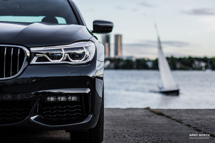 Black BMW 7 Series car by the water with a sailboat