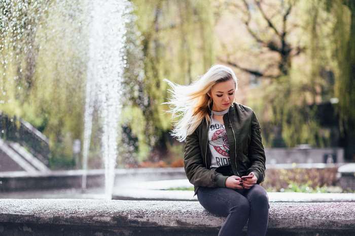 Young woman with blonde hair using phone near fountain