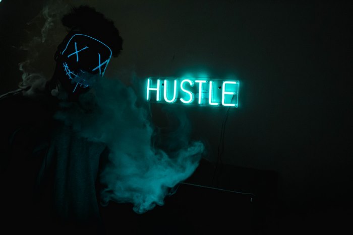 Person in a neon mask with 'Hustle' neon sign